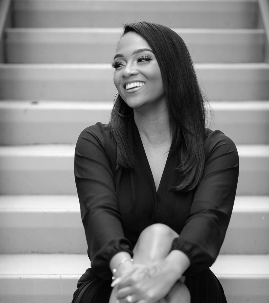 Professional portrait of Jessica Hester, a woman with long straight dark hair, smiling warmly while looking slightly off-camera, wearing large hoop earrings and a dark V-neck top, seated with arms crossed against a background of light-colored steps, in black and white