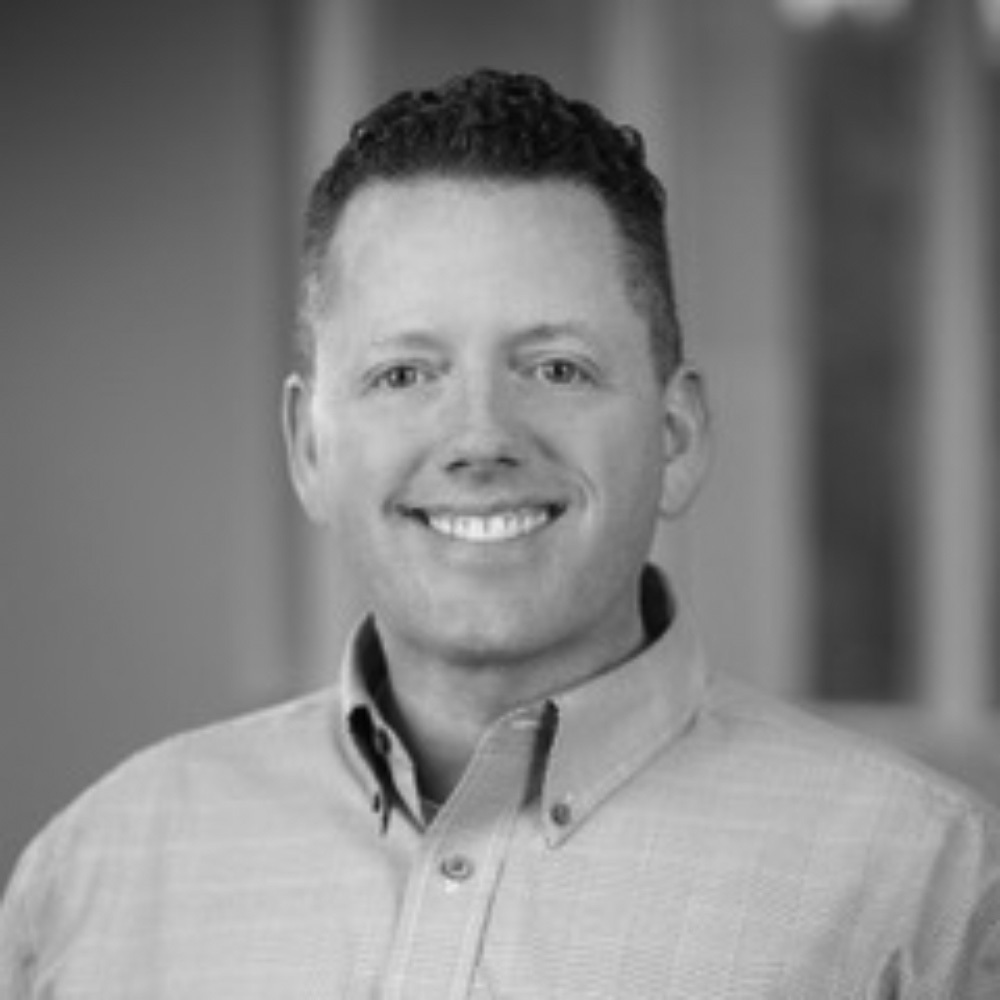 Professional headshot of Drew Glenn, a man with short curly hair, smiling confidently at the camera, wearing a button-down collared shirt against a blurred neutral indoor background, in black and white