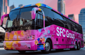 Colorful pink-dominant tour bus with vibrant multicolored bubble pattern wrap, "SFC Group" in large white letters on side, parked in city with tall buildings and pinkish sunset sky.